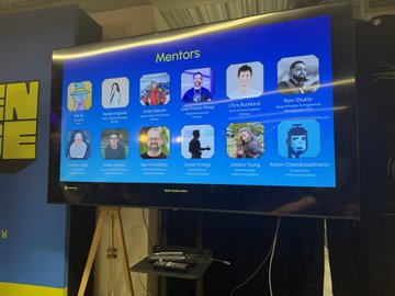 A television monitor on a stand displays a 'Mentors' slide featuring headshots and names of various industry professionals at a conference or event.