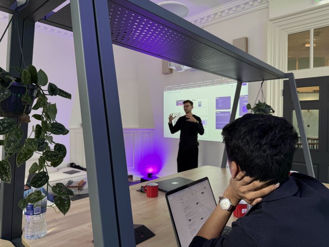 A meeting or presentation taking place in a modern room, where a man stands in front of a projected slide while another person sits at a table with a laptop, framed by a dark metal structure with a hanging plant.