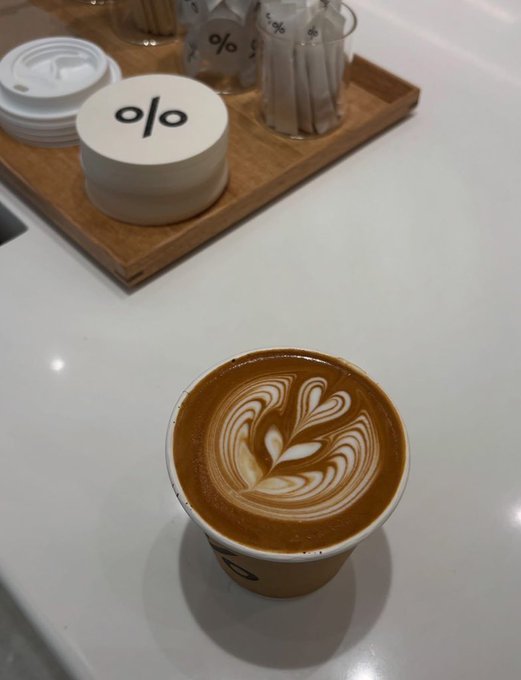 A latte with heart-shaped foam art in a '%' branded cup, sitting on a white counter next to a tray of coffee accessories.