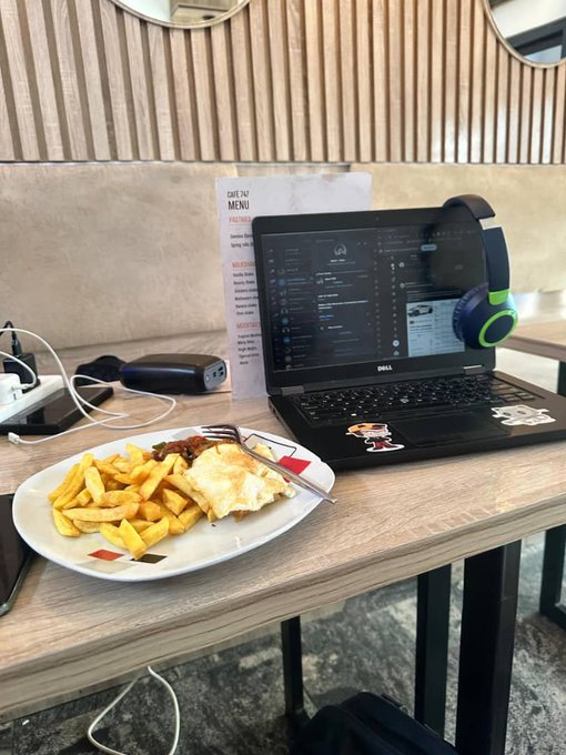 A laptop sits on a wooden table next to a plate of food with fries and a folded omelet, alongside headphones and a power bank at what appears to be a cafe.