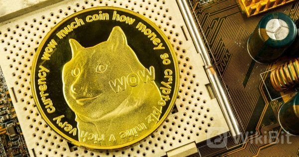 A Dogecoin physical coin resting on a computer motherboard, featuring the Shiba Inu dog and the text 'wow much coin how money so crypto very currency mine A' around the edge, with a WikiBit watermark.