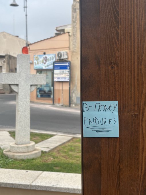A blue sticky note on a wooden door reads 'B-MONEY ENDURES', with a stone cross and street scene visible through a window.