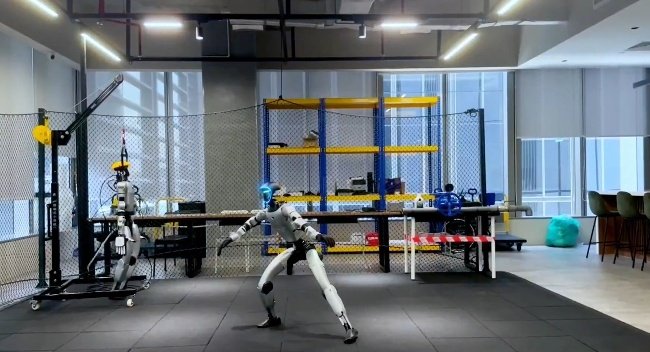 A humanoid robot in a motion-capture or training facility, posed dynamically with one leg lifted and arms extended, with a second robot visible in the background behind a safety net.