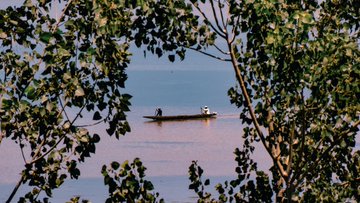 A serene photograph of a small boat with two figures floating on calm water, framed by tree branches with green leaves in the foreground.