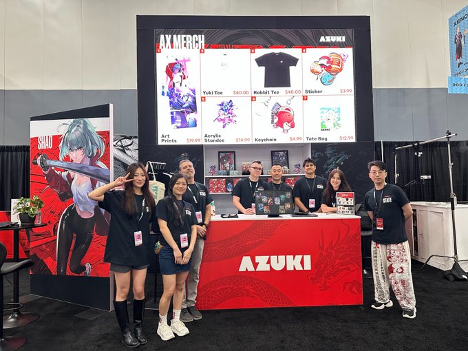 The Azuki merch booth at Anime Expo features a team of staff posing in front of a display showcasing various items including Yuki Tees ($40), Rabbit Tees ($40), Stickers ($3.99), Art Prints ($19.99), Acrylic Standees ($14.99), Keychains ($4.99), and Tote Bags ($59.99), with anime character banners promoting "SHAO" nearby. The image illustrates the post's message by showing the real people behind the brand, celebrating the team whose collective effort brings the Azuki project to life at conventions.
