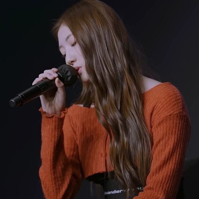 A young woman with long wavy brown hair wearing an orange ribbed sweater sings into a microphone with her eyes closed.