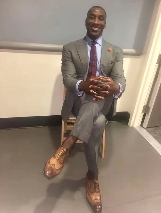 A smiling man in a gray suit, blue shirt, and patterned tie sits on a chair with his legs crossed, wearing distinctive two-tone brown leather shoes.