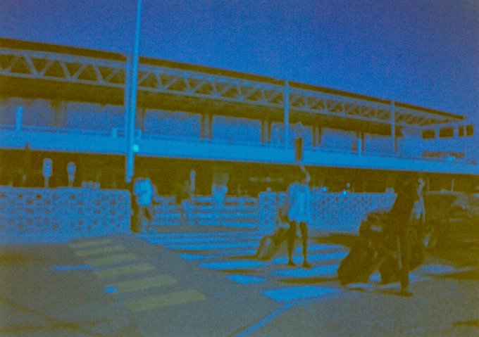 A lone figure stands in front of a large stadium structure under an early morning sky, carrying bags and appearing to arrive or depart at dawn. The blue-tinted, grainy quality of the scene captures that quiet, groggy feeling of being up before the world feels fully awake.