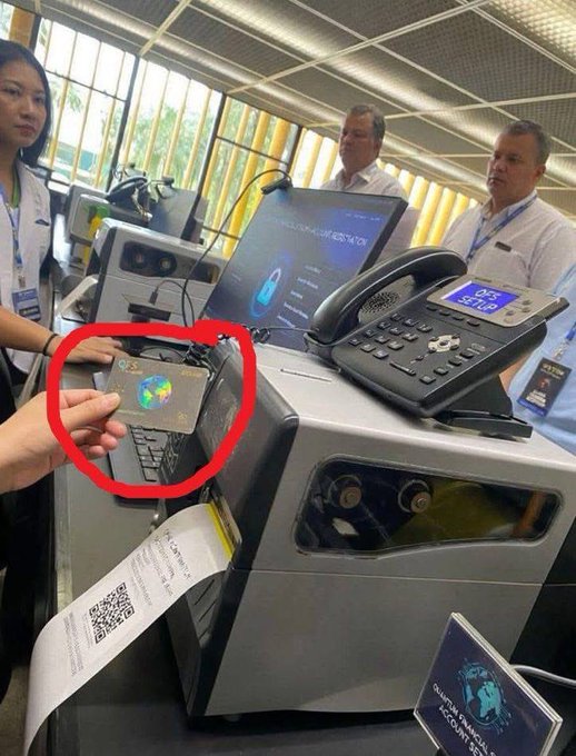 A person's hand holds up a dark card featuring a holographic Earth globe design, circled in red, next to what appears to be a passport/border control station with "DEPT" visible on a telephone display. The image relates to the post by showing the physical "QFS card" being demonstrated at what looks like a border control or check-in counter.