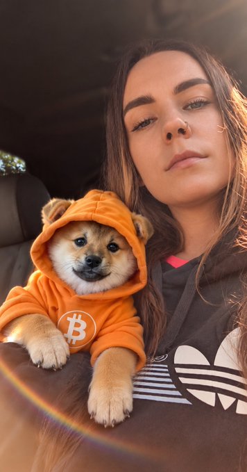 A selfie of a young woman with a nose ring posing with a small dog wearing an orange Bitcoin-themed hoodie.
