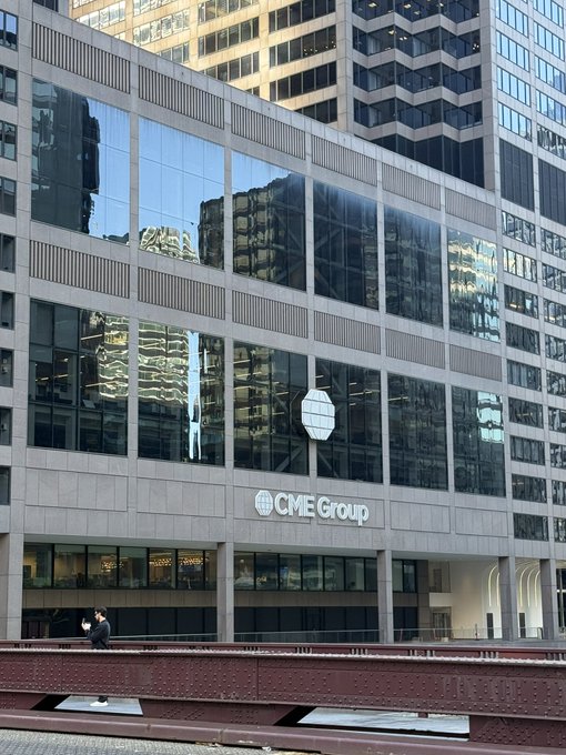 The exterior of a large building is shown with a 'CME Group' logo and name displayed prominently on the facade above ground-level windows, with tall office towers visible in the background.