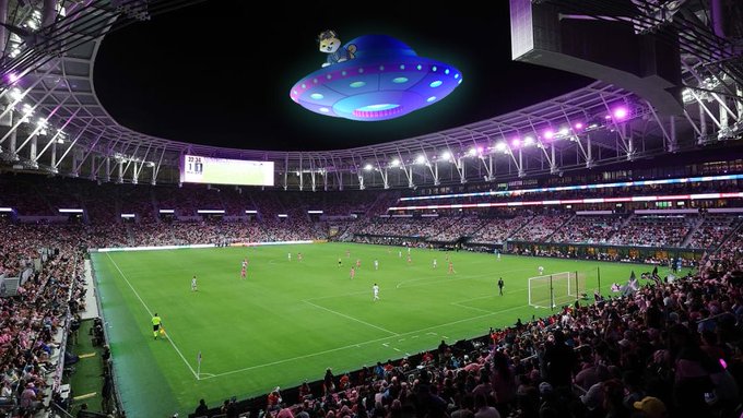 The image shows a large stadium filled with people watching a soccer game between Brazil and Scotland. A purple and blue UFO is hovering above the field, capturing the attention of the spectators. The crowd appears to be engaged in the game, while some are also looking at the UFO. The photo has been edited to include a caption that reads "TRENDING: TODAY IS THE DAY! A Brazilian psychic claims a UFO will appear above Hard Rock Stadium during Brazil vs Scotland at the 2026 World Cup on June 24." This post seems to be related to an event or situation involving the UFO, possibly drawing attention from fans and players alike.