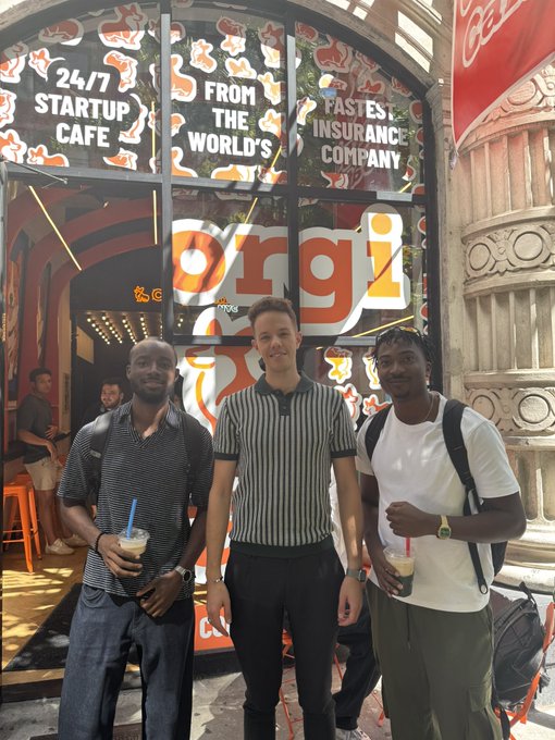 Three men posing for a photo with iced coffees outside an 'Orgi' storefront, which has window graphics reading '24/7 STARTUP CAFE' and 'FROM THE WORLD'S FASTEST INSURANCE COMPANY'.
