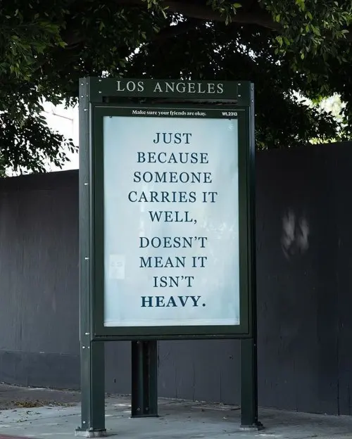 A street sign in Los Angeles displaying the message 'JUST BECAUSE SOMEONE CARRIES IT WELL, DOESN'T MEAN IT ISN'T HEAVY.' with the smaller text 'Make sure your friends are okay. WL200' above it.