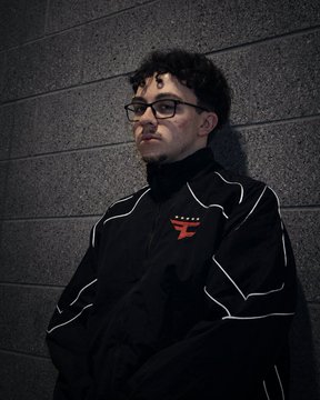 A young man with curly dark hair and glasses, wearing a black jacket with white piping, stands against a textured gray concrete wall.