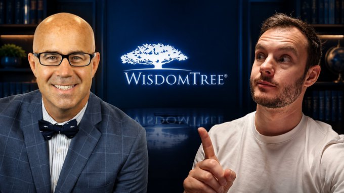 Two men pose in front of a WisdomTree logo backdrop, with one man on the left wearing glasses and a bow tie smiling at the camera, while the other on the right in a white shirt points with a serious expression.