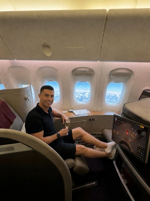 Cristiano Ronaldo relaxing in what appears to be a first-class airplane seat (Qatar Airways) with a glass of champagne and a screen displaying a flight map.