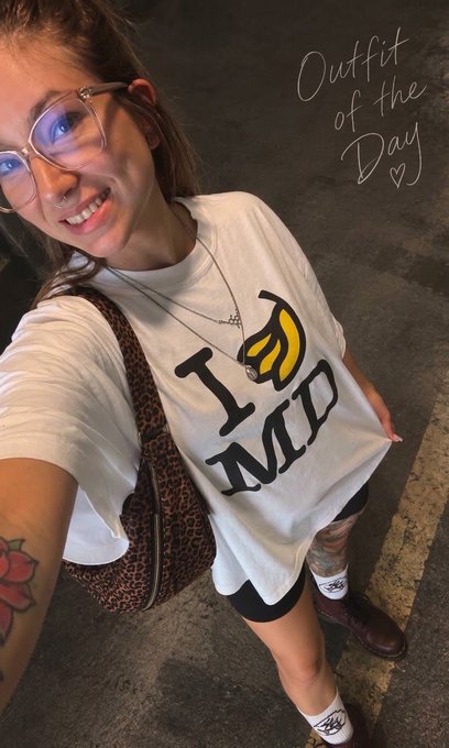 A selfie of a smiling woman wearing glasses, a white t-shirt that reads 'I [banana] MC', a leopard print bag, with the handwritten text 'Outfit of the Day ♡' in the upper right corner.