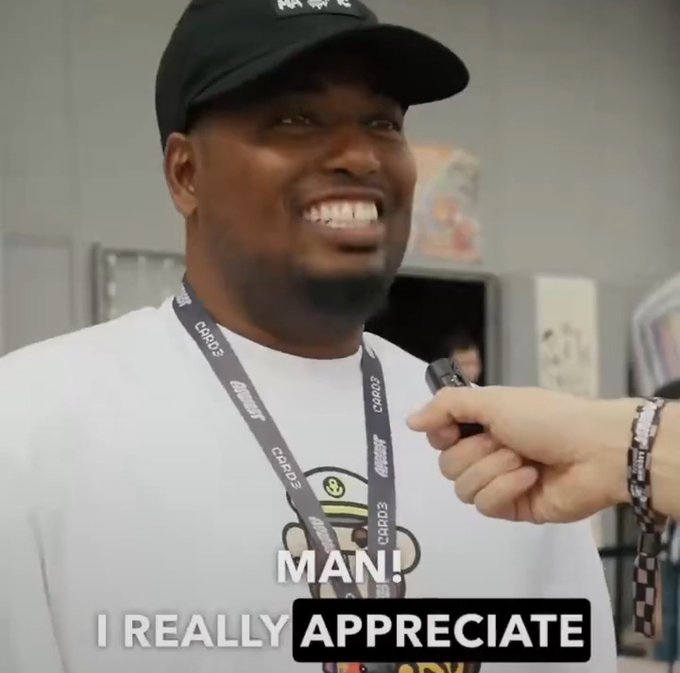 A man wearing a black cap and white t-shirt with a lanyard labeled "C4R13" smiles widely in an interview setting, with subtitles reading "MAN! I REALLY APPRECIATE".