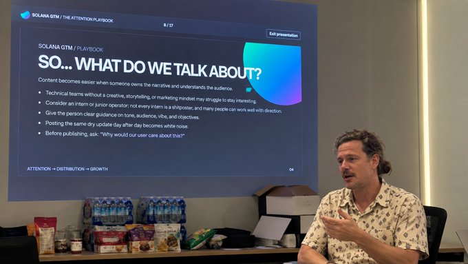 A man with long hair speaking in front of a presentation slide titled 'SO... WHAT DO WE TALK ABOUT?' under the header 'SOLANA GTM / PLAYBOOK,' with snacks and water bottles arranged on a table in front.