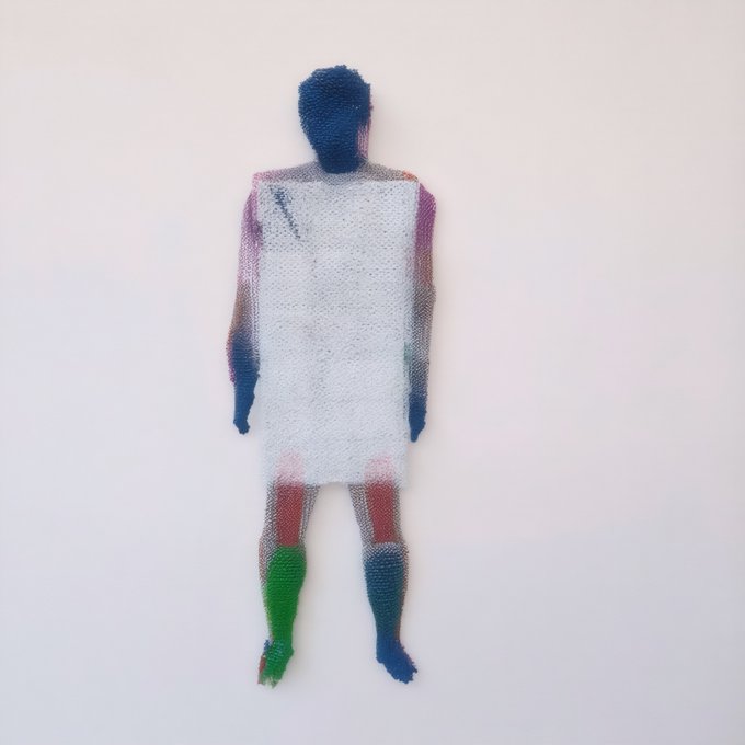 A textile artwork or sculpture depicts a human figure made of white mesh or netting over a colorful, multicolored body, mounted on a white wall.