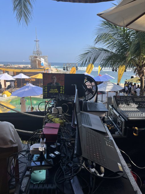 A beachside DJ or event setup with laptops, a mixing console, and tangled cables in the foreground, overlooking an ocean venue with umbrellas, palm trees, and a stage featuring a 'CLUB HOUSE' sign.