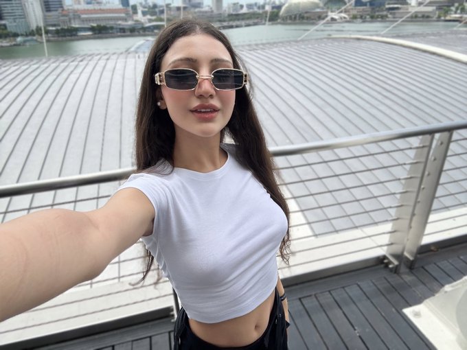 A woman wearing rectangular sunglasses and a white crop top takes a selfie on a balcony overlooking a cityscape with a distinctive dome-shaped roof and waterfront.