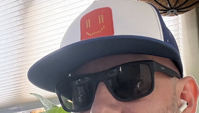A selfie of a person wearing a white and navy cap with a red smiling face patch and dark sunglasses, with an earbud visible.