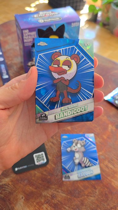 A hand holding a Topps Chrome Animal Crossing trading card of 'Bandicoot' with another 'Raccoon' card on the table, alongside a Real Chrome booster box featuring Sanrio characters.