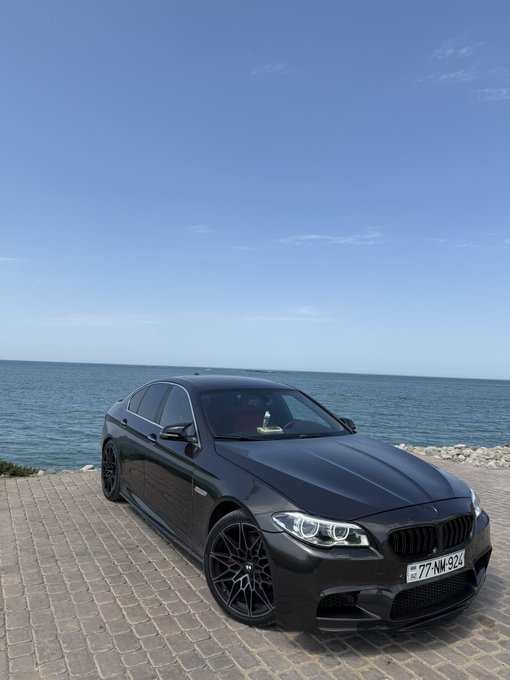 A matte black BMW sedan parked on a brick pathway beside the ocean under a clear blue sky, displaying a license plate reading '77-NM-924'.