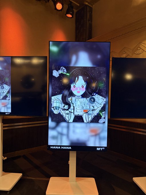 A digital display showcasing an NFT artwork titled "HANA HANA" featuring a stylized illustration of a girl with space-themed elements on her chest, displayed on a stand in what appears to be a gallery setting.