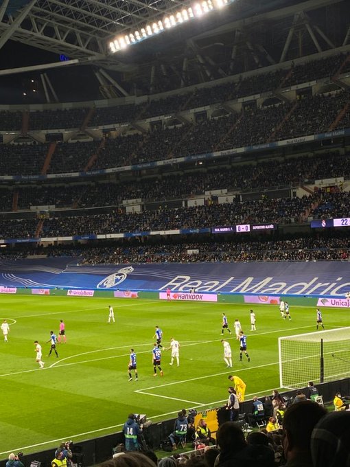 A nighttime view of a Real Madrid football match at the Santiago Bernabéu Stadium, with players in white on the pitch and the scoreboard showing Real Madrid 0-0, with the home crowd filling the stands.
