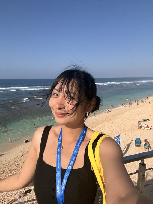 A selfie of a smiling young woman with dark hair and bangs, wearing a black top and a blue lanyard, taken at a sunny beach overlooking the ocean.