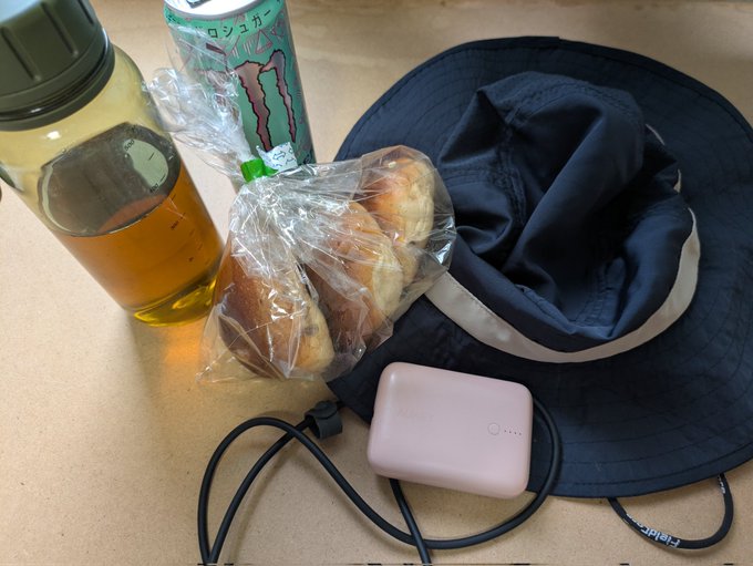 A pink portable charger, navy bucket hat, Monster energy drink, water bottle, and bag of bread buns laid out on the floor, likely packed as supplies for a 42km running challenge in STEPN.

Visible text includes "モンスター" (Monster) on the green can and "42km" referenced in the post about today's marathon attempt and revenge run past a 700m tunnel.