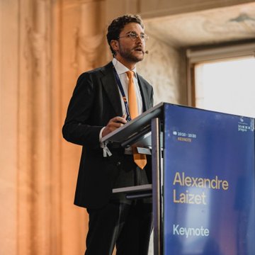 A man in a suit and orange tie gives a keynote presentation at a podium displaying "Alexandre Laizet Keynote" at a 2025-2026 event.
