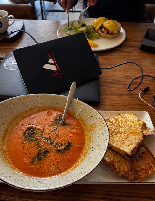 A meal at a café or restaurant featuring a bowl of tomato soup with herbs and grilled cheese sandwich on a wooden table, alongside a laptop, plate of eggs benedict, and coffee.