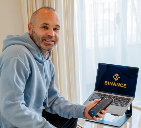 A man in a light blue hoodie sits smiling in front of a laptop displaying the Binance logo while holding a smartphone showing a trading app.