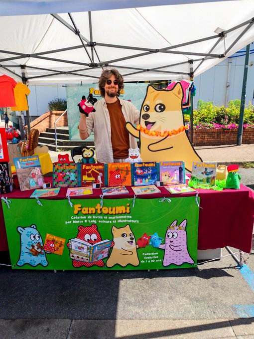A man stands under a white canopy at a book fair, smiling and holding a plush toy at his vendor booth for 'Fantoumi', a French children's book collection.