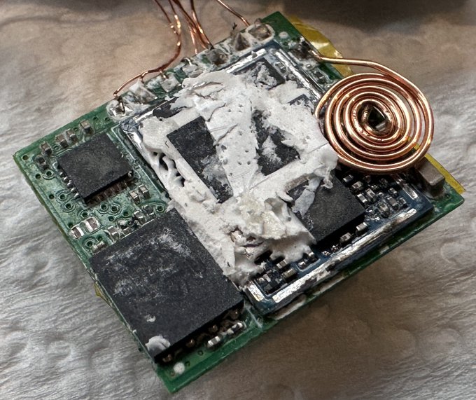 A teardown photo showing a Ledger Nano X hardware wallet's internals, with the secure element chip exposed after the epoxy potting compound was removed to reveal the implanted hardware, alongside the extracted wireless charging coil. The image accompanies a security research presentation revealing a hardware implant attack on the cryptocurrency wallet device.