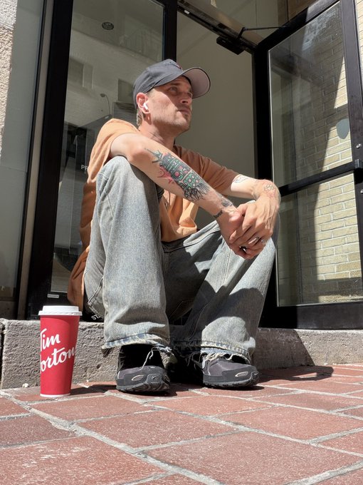 A photograph of a man wearing a baseball cap, orange shirt, and jeans sitting on a step outside a building, with a red Tim Hortons coffee cup placed on the brick ground beside him.