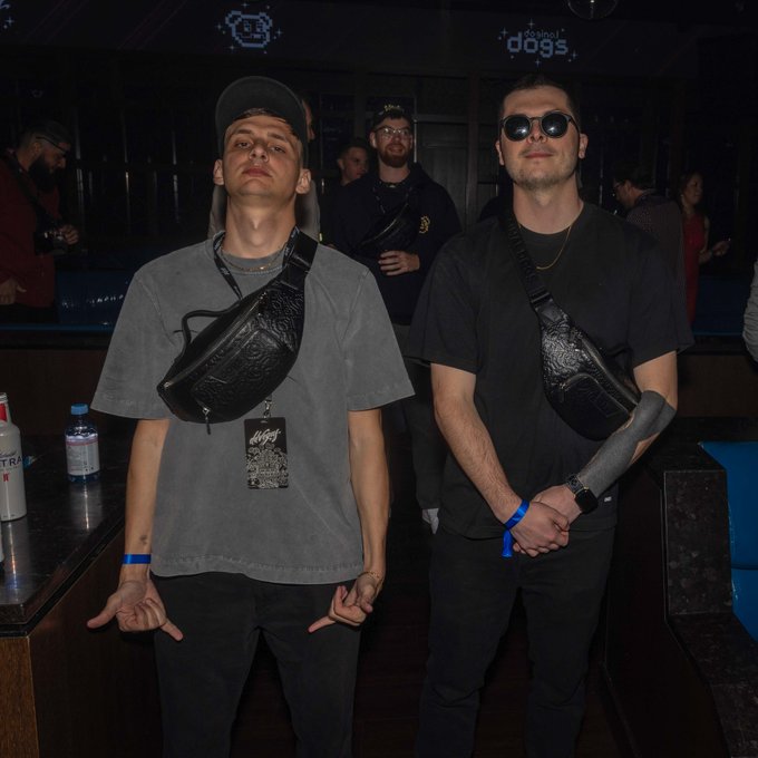 Two men pose at an event with 'School of Dogs' signage visible above; the man on the left wears a gray t-shirt and backwards cap with a black crossbody bag, while the man on the right wears sunglasses and a black shirt with a similar bag.