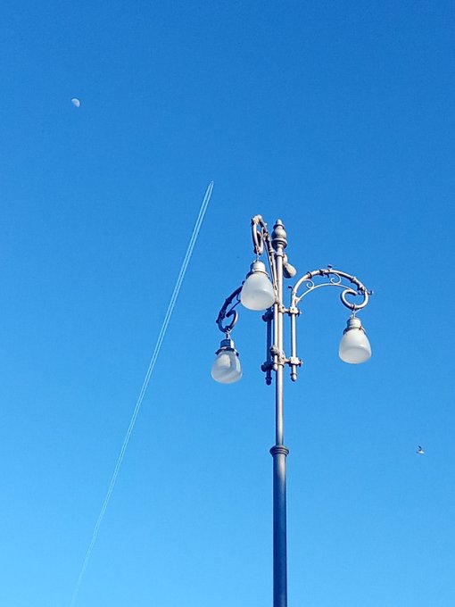 An ornate multi-globe street lamp silhouetted against a bright blue sky with the moon faintly visible in the upper left.