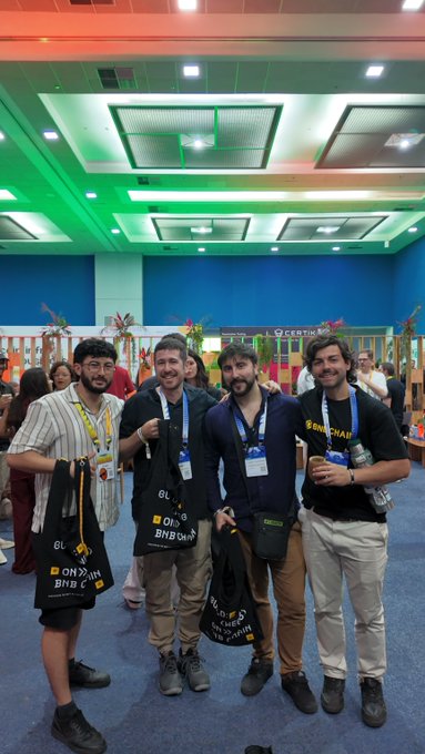 Four young men pose together at what appears to be a conference or event, with two holding bags labeled 'BNB CHAIN' and the rightmost wearing a shirt that says 'BNB CHAIN'.