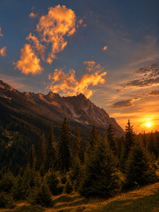 A scenic landscape photograph showing a sunset over a rugged mountain range with dense evergreen forests in the foreground and dramatic orange-lit clouds in the sky.
