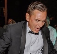 A man in a gray suit and bow tie looking down with a distressed expression, appearing to be at a crowded event.