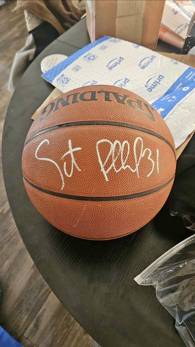 A Spalding basketball with "SJ2 PEREZ" signed in white marker sits on a dark wooden table alongside a blue Amazon Prime envelope. The image supports the post's humor about an underpriced $80 signed basketball, suggesting it's actually worth significantly more as an autographed collectible.