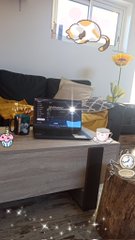 A living room scene featuring a wooden table with a laptop, teacup, and a vase with a yellow tulip, with sunlight streaming through a window adorned with stained glass.
