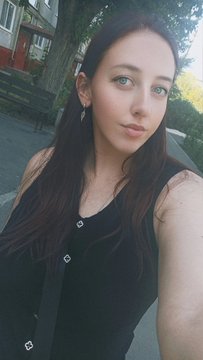 A selfie of a young woman with long dark hair and green eyes, wearing a black top and earrings, taken outdoors with greenery in the background.