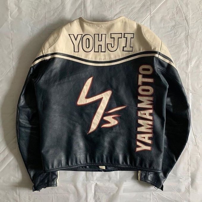 A black leather Yohji Yamamoto jacket laid flat, featuring 'YOHJI' across the shoulders, 'YAMAMOTO' down one side, and a large Y's logo on the back, with cream-colored shoulder panels.