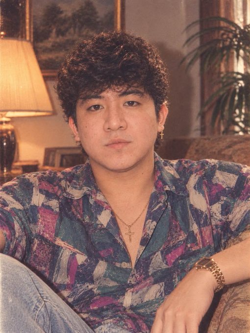 A man with curly hair wearing a colorful patterned shirt and gold cross necklace sits on a couch in a retro-styled room.